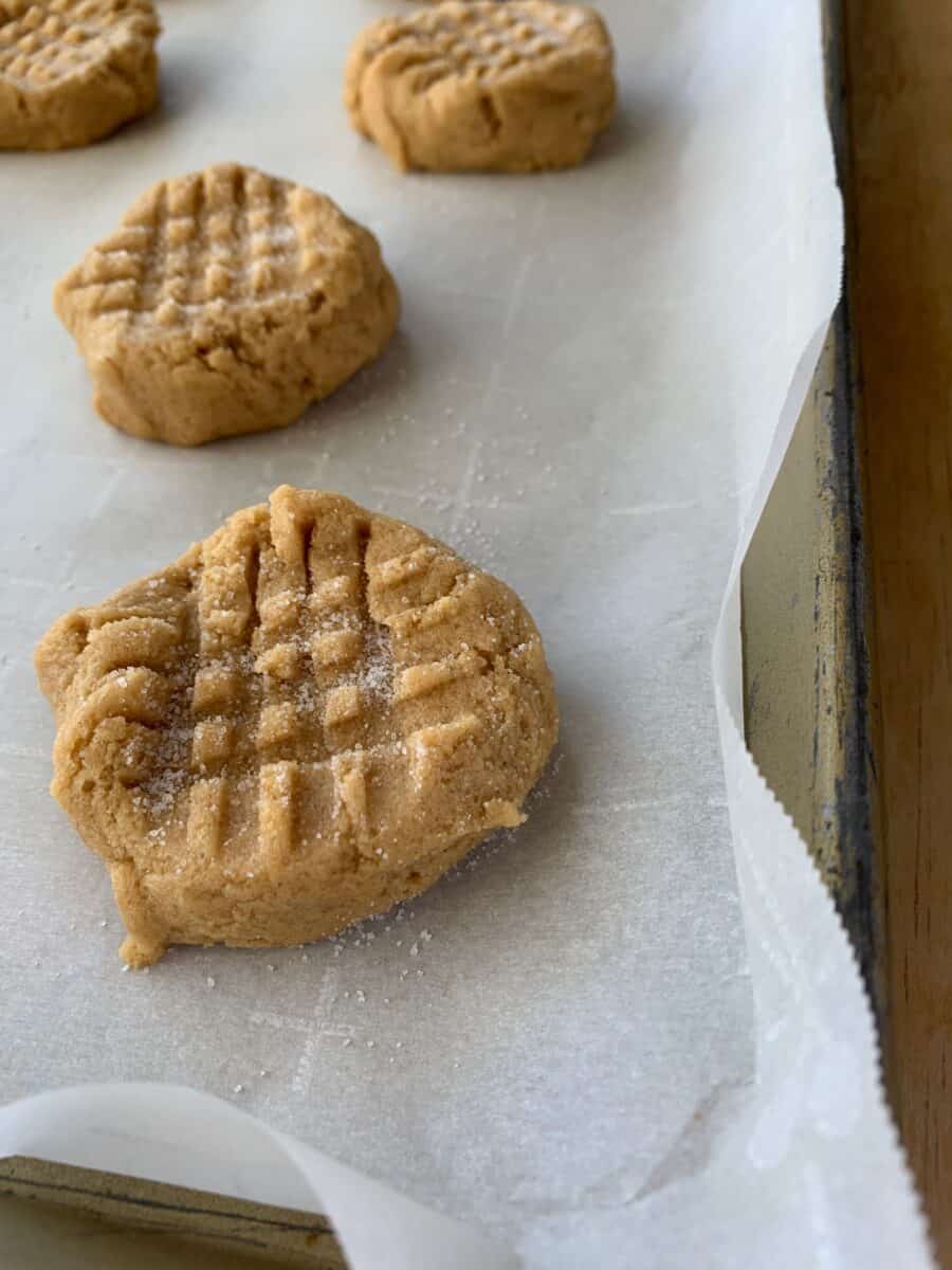 Peanut butter cookies on a baking sheet, with a criss cross pattern in the dough. 