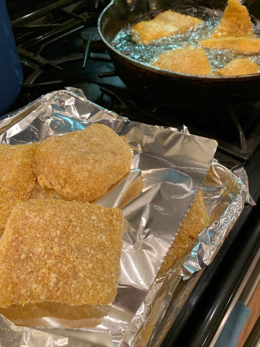 Breaded cot on foil next to a skillet, and several pieces of fish cooking in the skillet. 