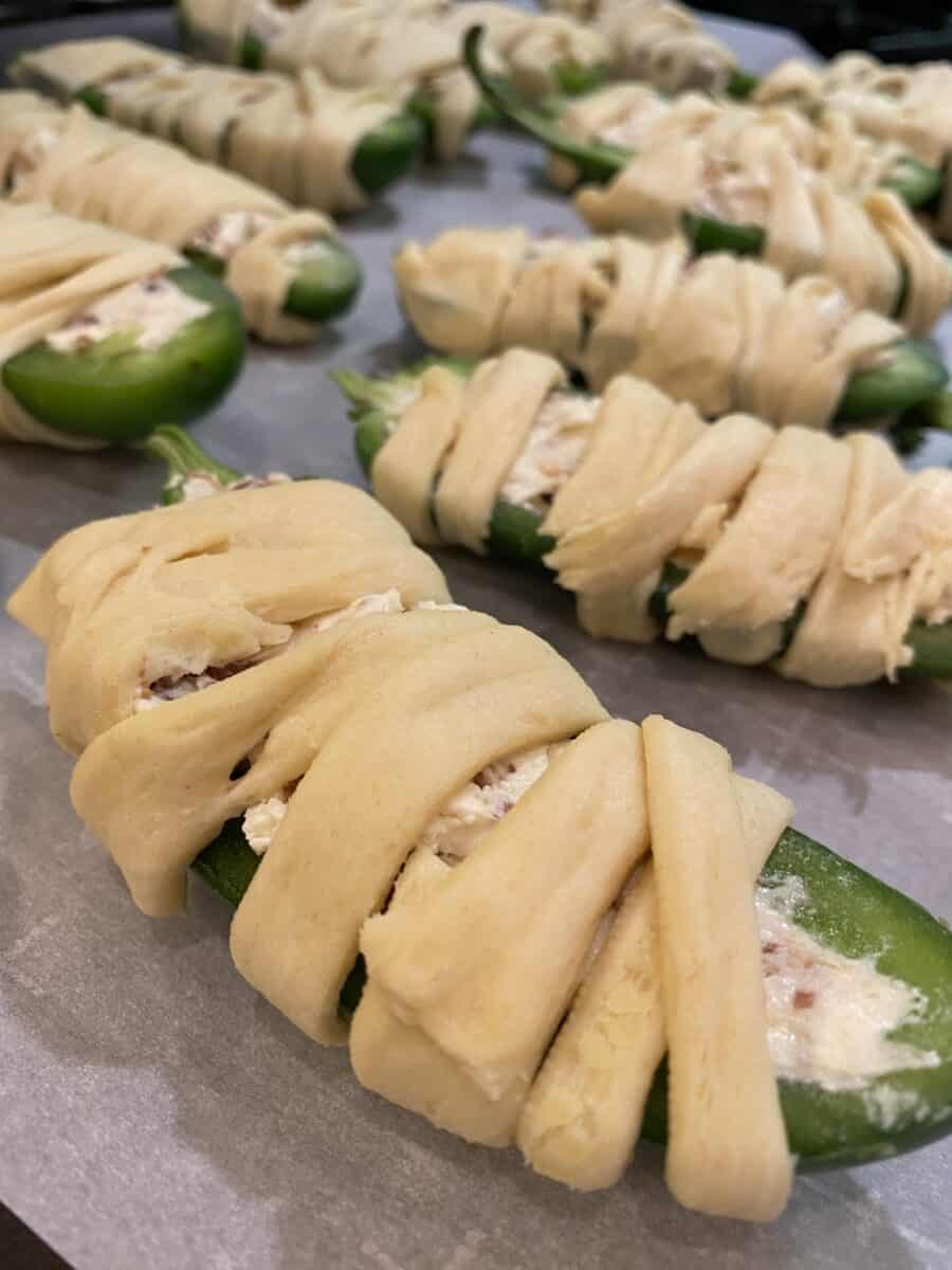 Unbaked jalapeno halves wrapped in crescent dough on a baking sheet. 