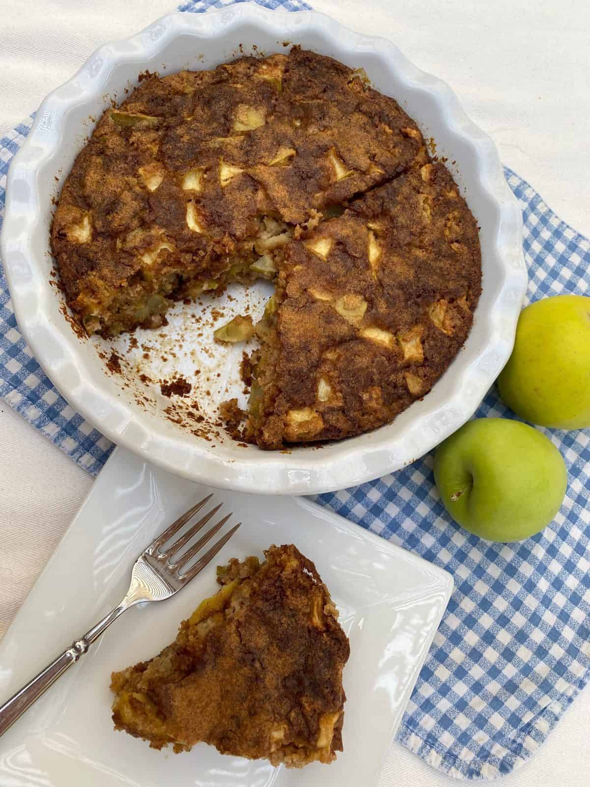 A wedge of apple cake with the baking dish and and apples in the background.