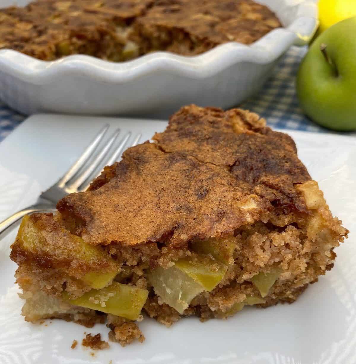 A wedge of apple cake with the baking dish and an apple in the background.