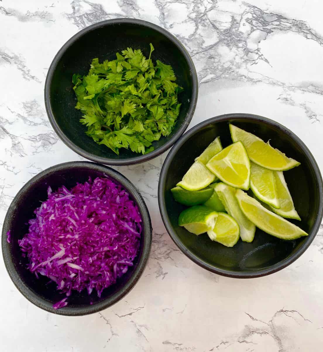 Red cabbage, limes and cilantro in small bowls on a counter.