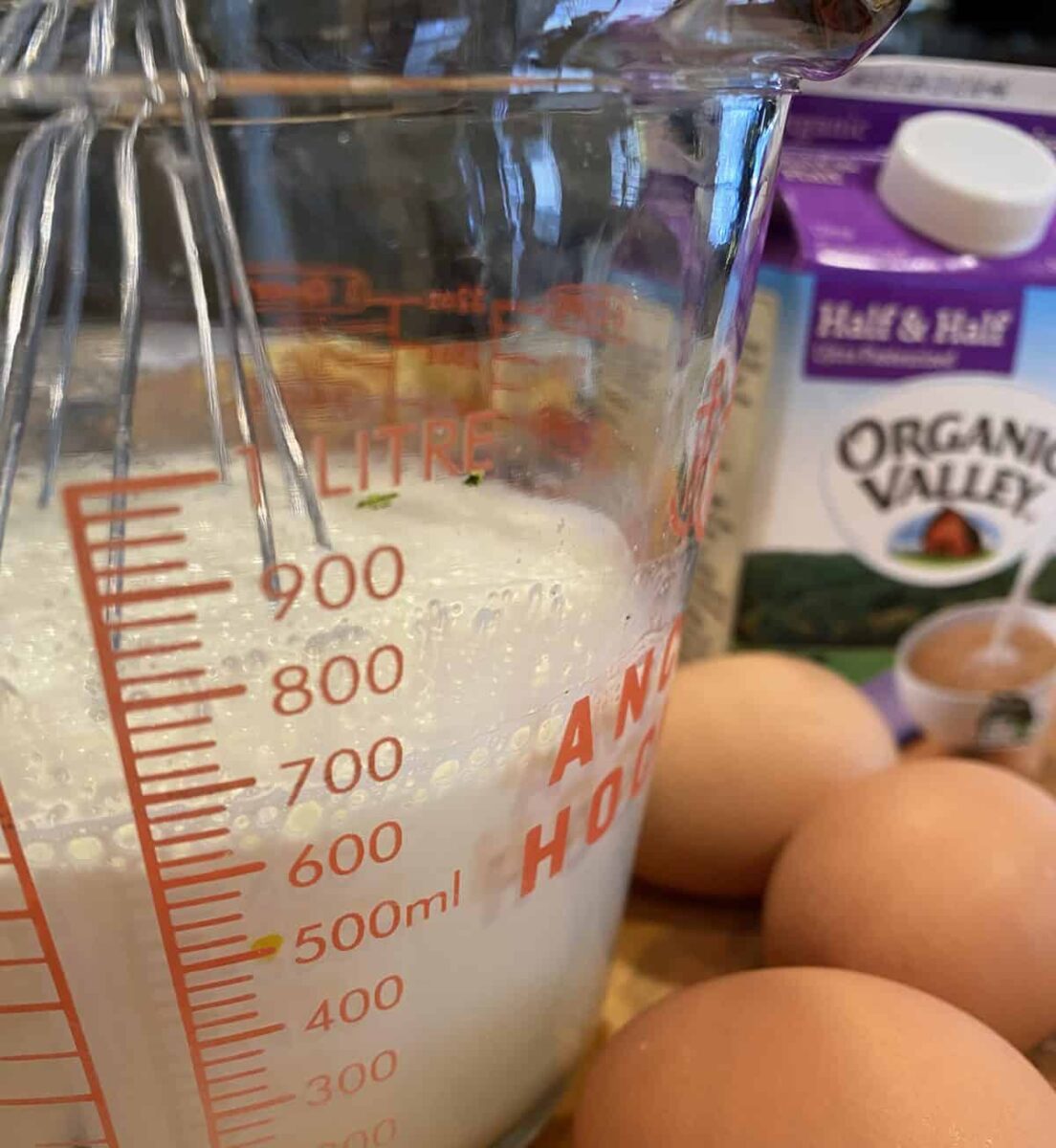 Half and half in a measuring cup with a whisk and three eggs.