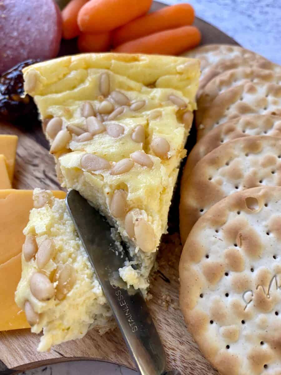 A wedge of savory cheesecake with crackers, cheddar cheese, carrots and a small knife on a cutting board.