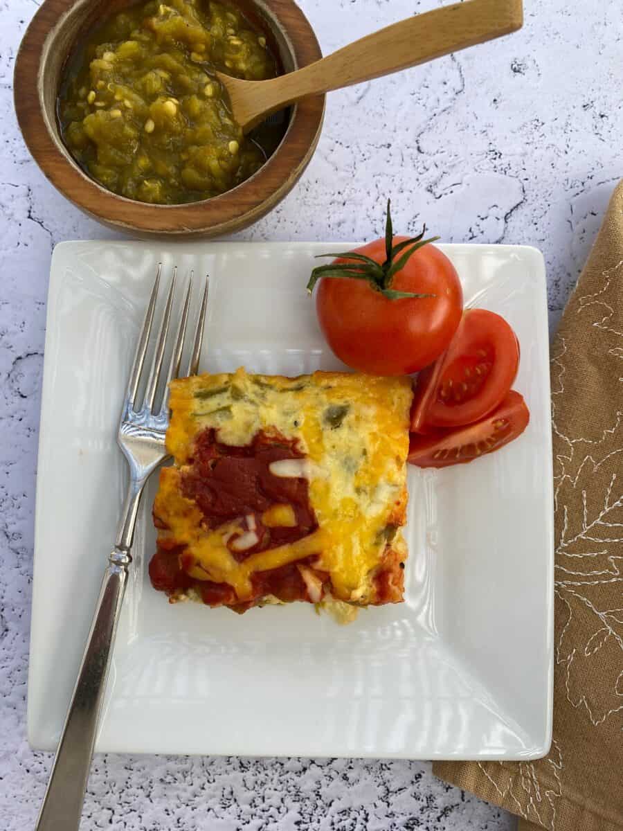 A plate with casserole, tomatoes and a fork. A bowl of salsa and a napkin on the side.