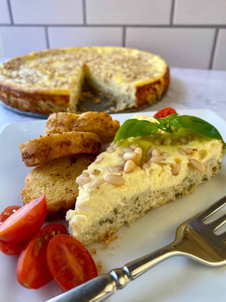 Slice of savory cheesecake with cherry tomato slices, small bread slices and a fork on the side. Whole cheesecake in the background.