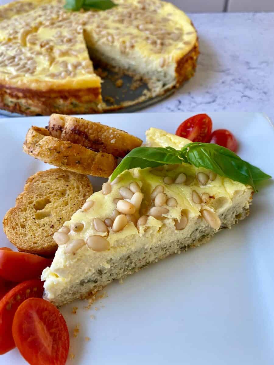 A wedge of cheesecake with cherry tomato slices and small rounds of bread. The whole cheesecake is in the background.