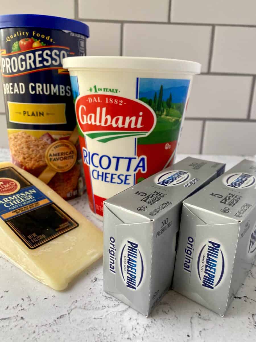 Cheesecake ingredients on a counter, cream cheese, parmesan cheese, ricotta cheese and a box of dry breadcrumbs.