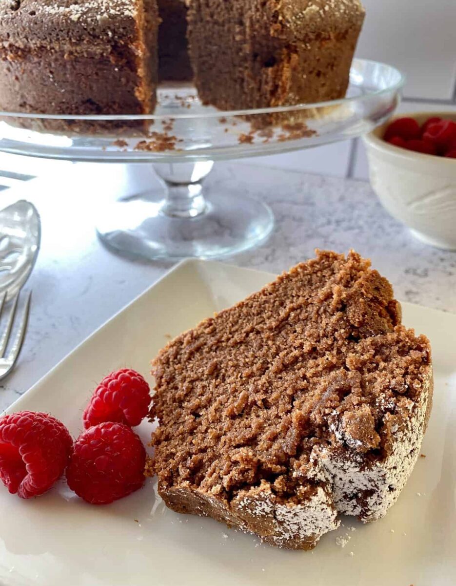 A slice of cake with raspberries with the entire cake on a serving plate in the background.