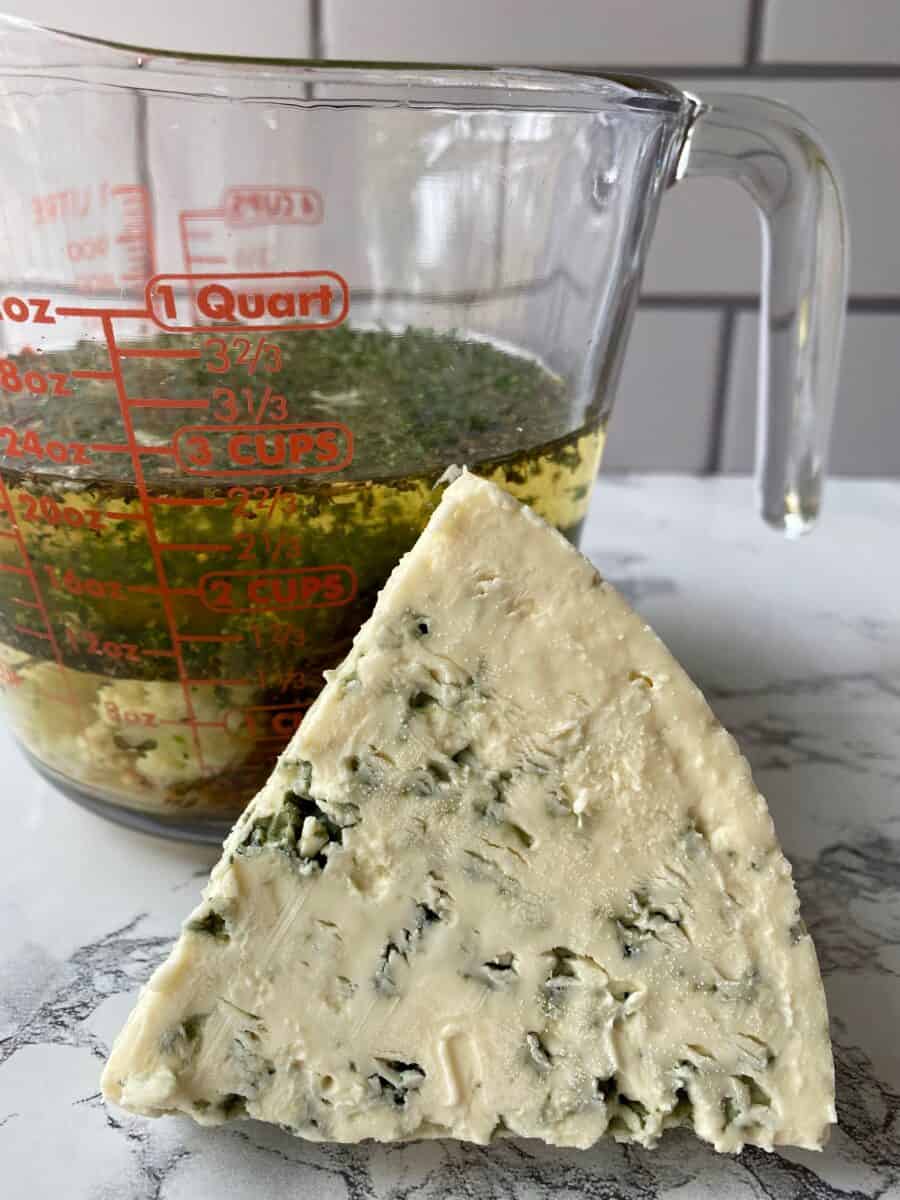 A wedge of blue cheese tipped on edge next to a glass measuring cup of salad dressing ingredients.