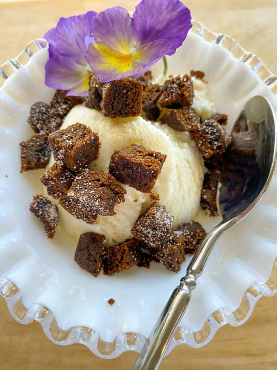 A scoop of ice cream with diced molasses snap on top and a pansy flower and spoon.