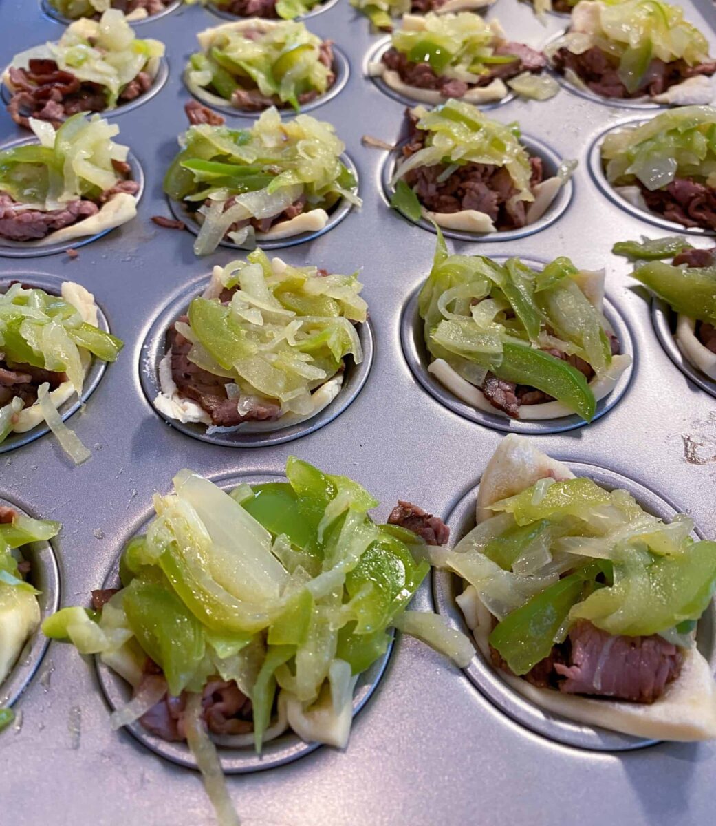 Mini Philly Cheesesteaks in a mini-muffin pan with cooked veggies on top of each one.