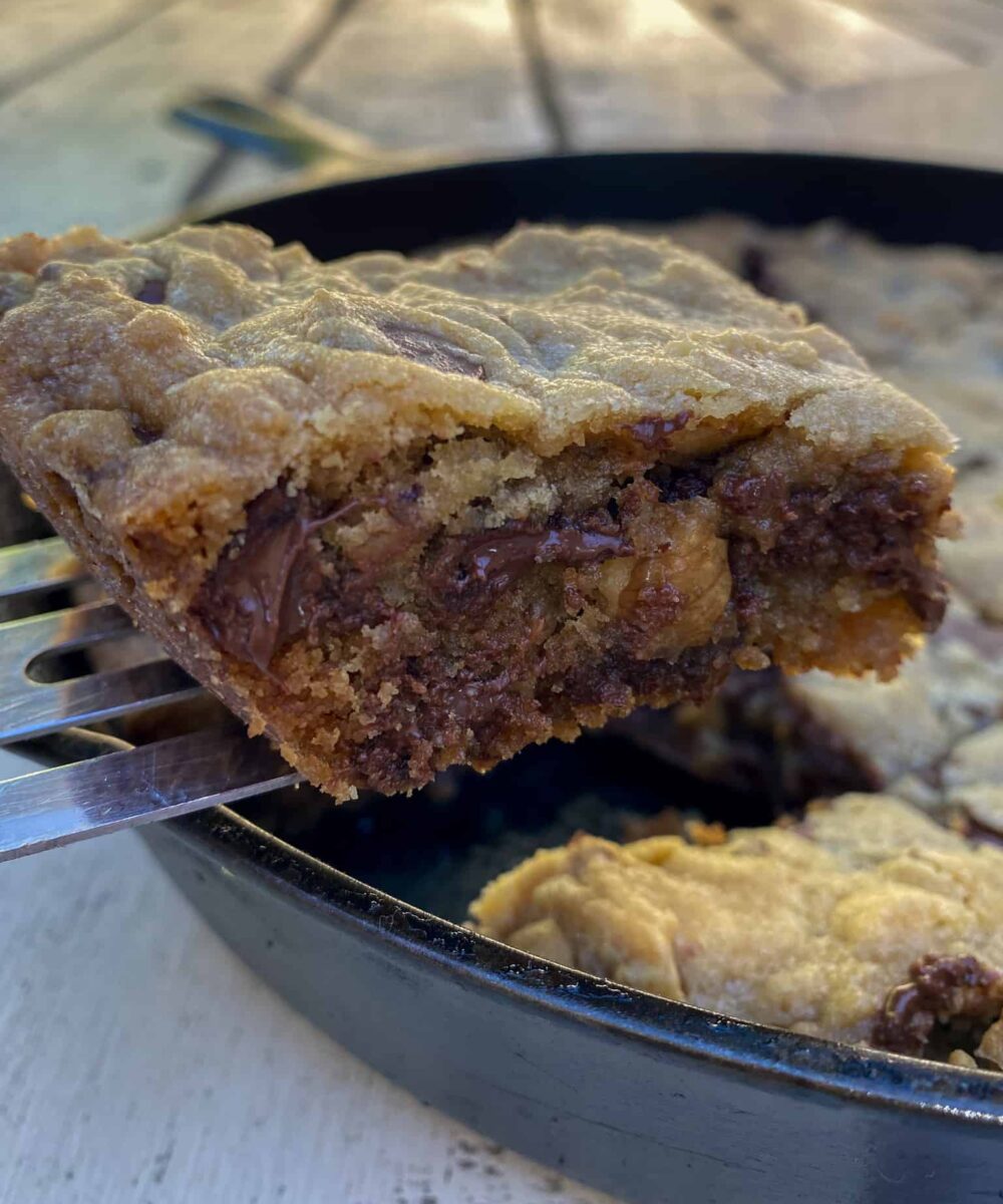 Chocolate chip bar cookie on a spatula, lifted out of a cast iron skillet on an outdoor table.