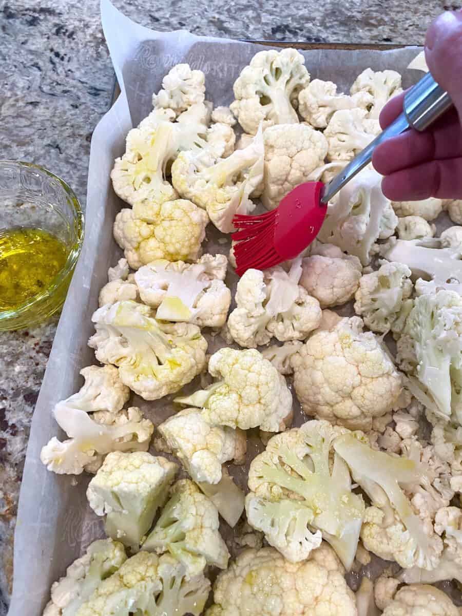 Raw cauliflower pieces on a baking pan being brushed with olive oil. 