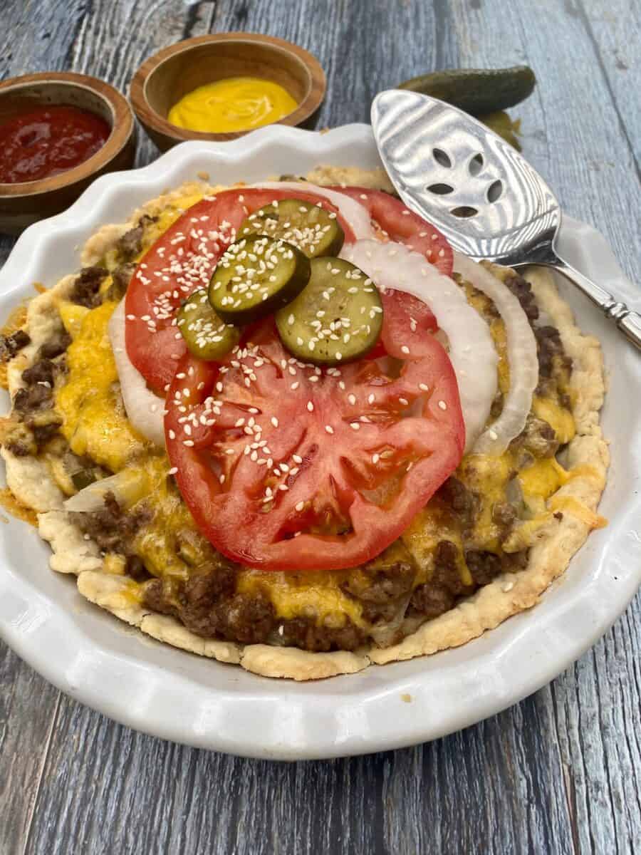 Cheeseburger pie in a pie plate with tomato and onion and sliced pickle on wooden planks