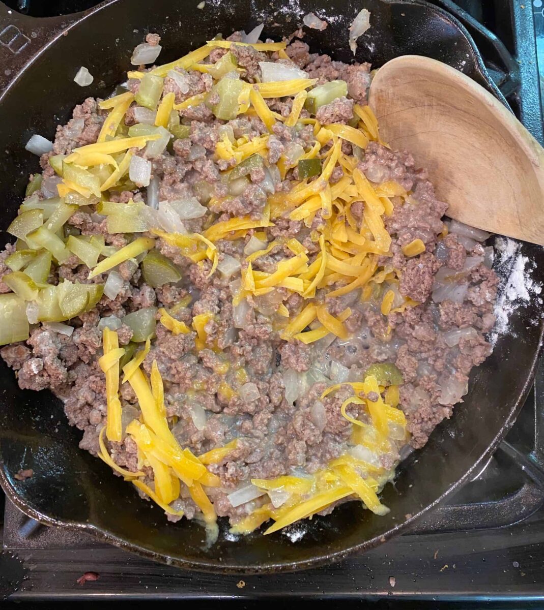 Fried hamburger with sliced cheese diced pickles in a cast iron skillet on a stove.