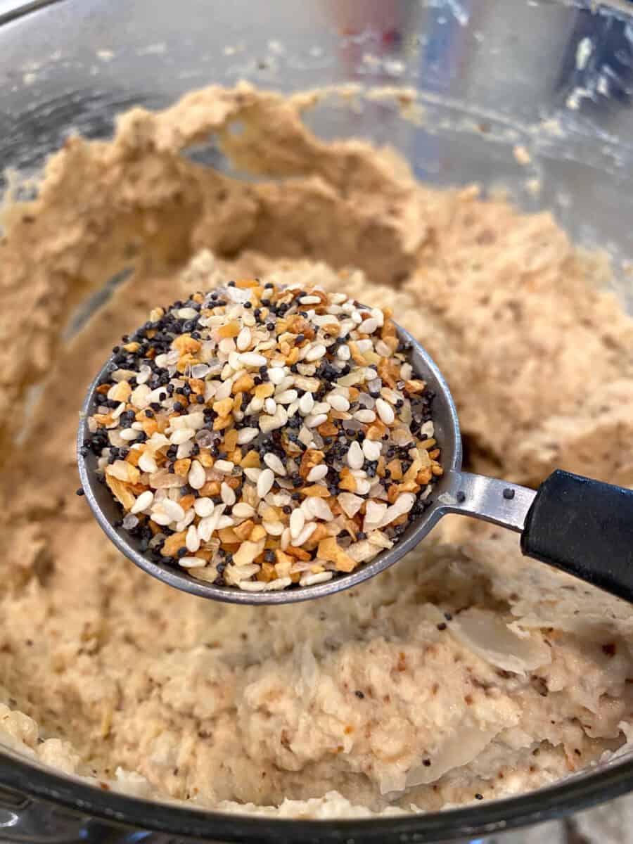 Everything Bagel seasoning on a tablespoon held over the blended cauliflower hummus.