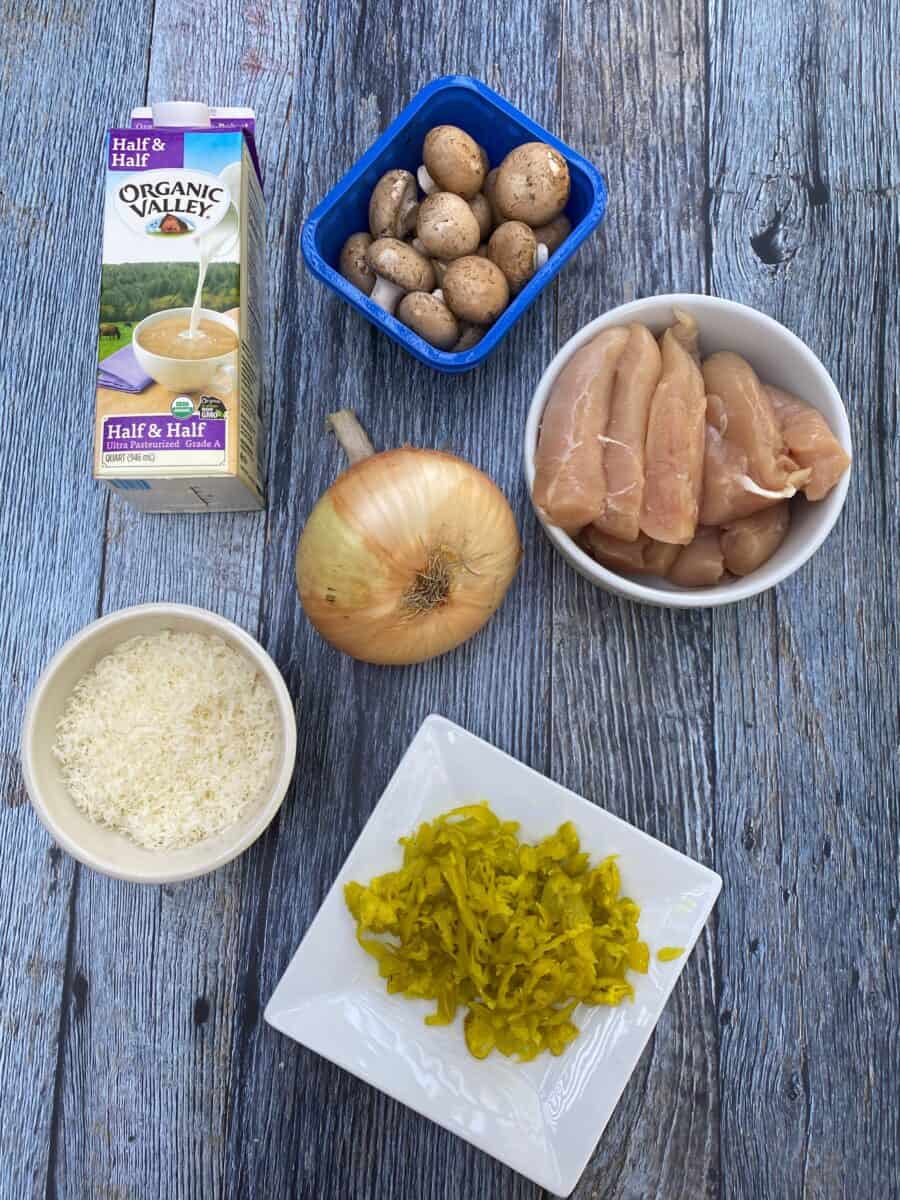 Spicy Chicken Alfredo ingredients on a wooden block, displaying a carton of half and half, mushrooms, uncooked chicken, a sweet onion, grated parmesan cheese and a plate of pepperoncinis.