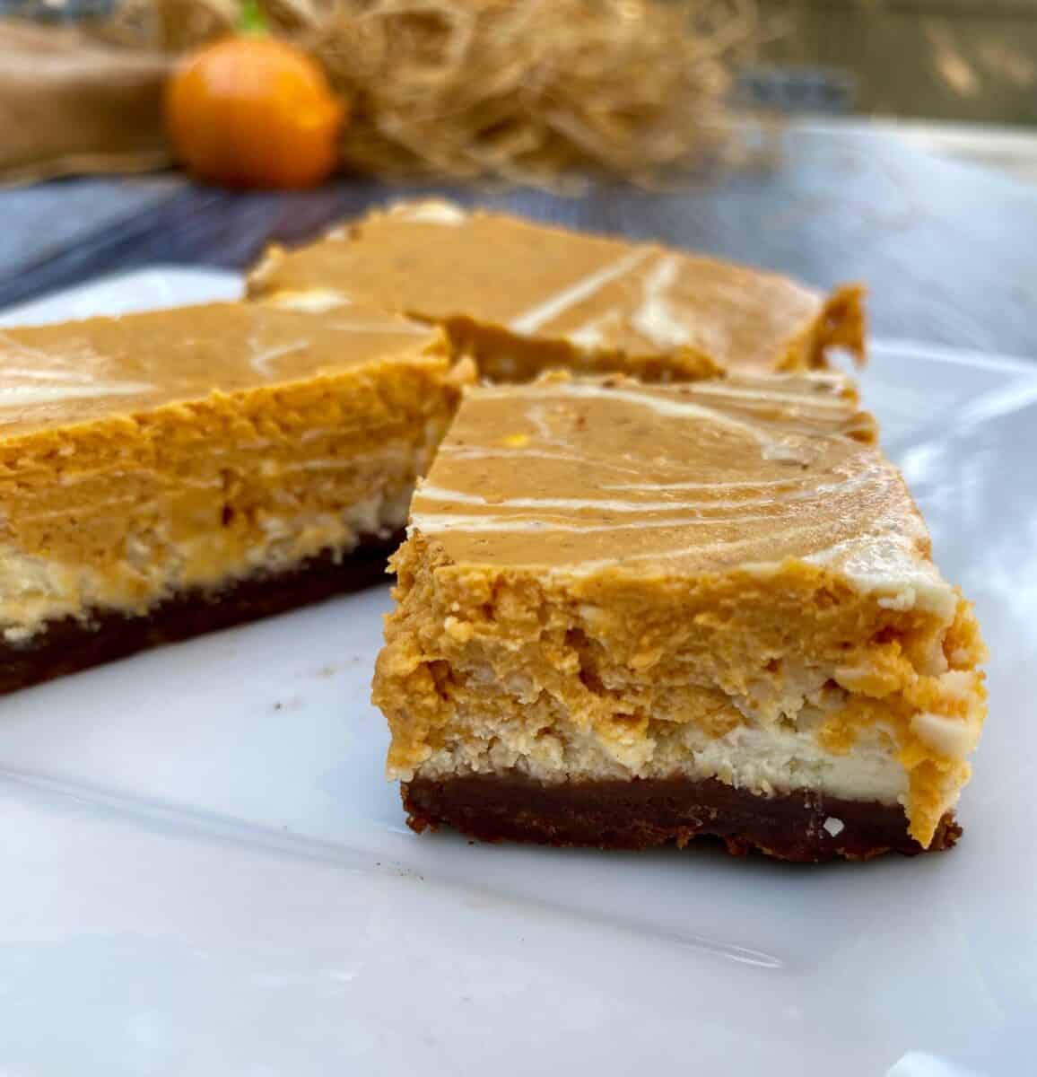 Three portions of pumpkin cheesecake bars on a plate with a small pumpkin and straw in the background.