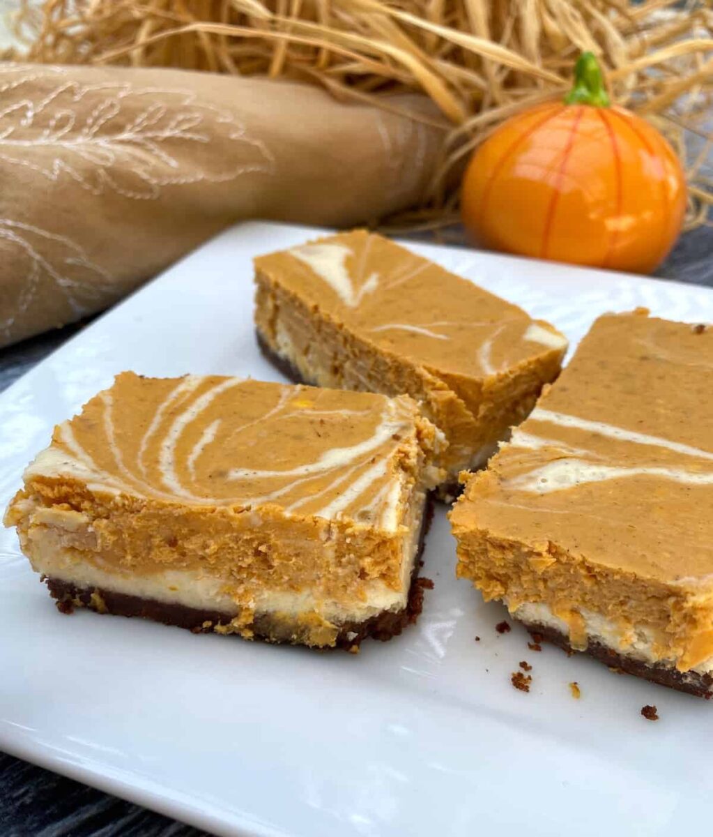 Three pumpkin cheesecake bars on a plate with straw and a miniature pumpkin ornament in the background.