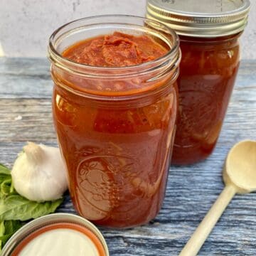 Sunday gravy in jars with a lid and a wooden spoon on the side next to a bulb of garlic and fresh basil