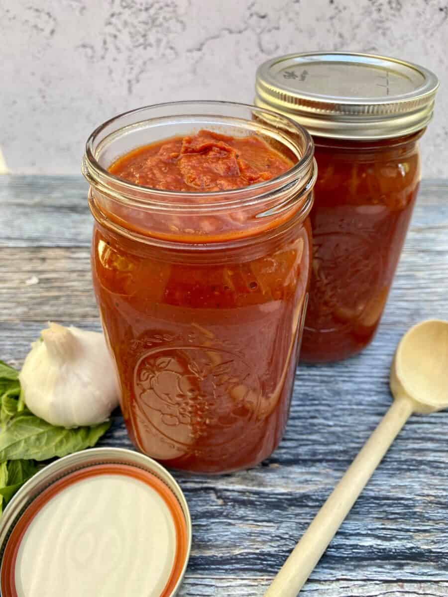 Sunday gravy in jars with a lid and a wooden spoon on the side next to a bulb of garlic and fresh basil