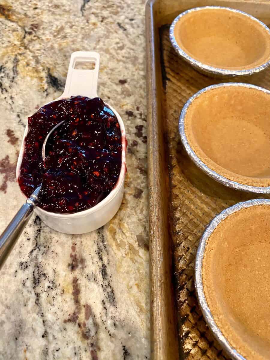 A half cup of jam next to a baking sheet filled with mini pie crusts