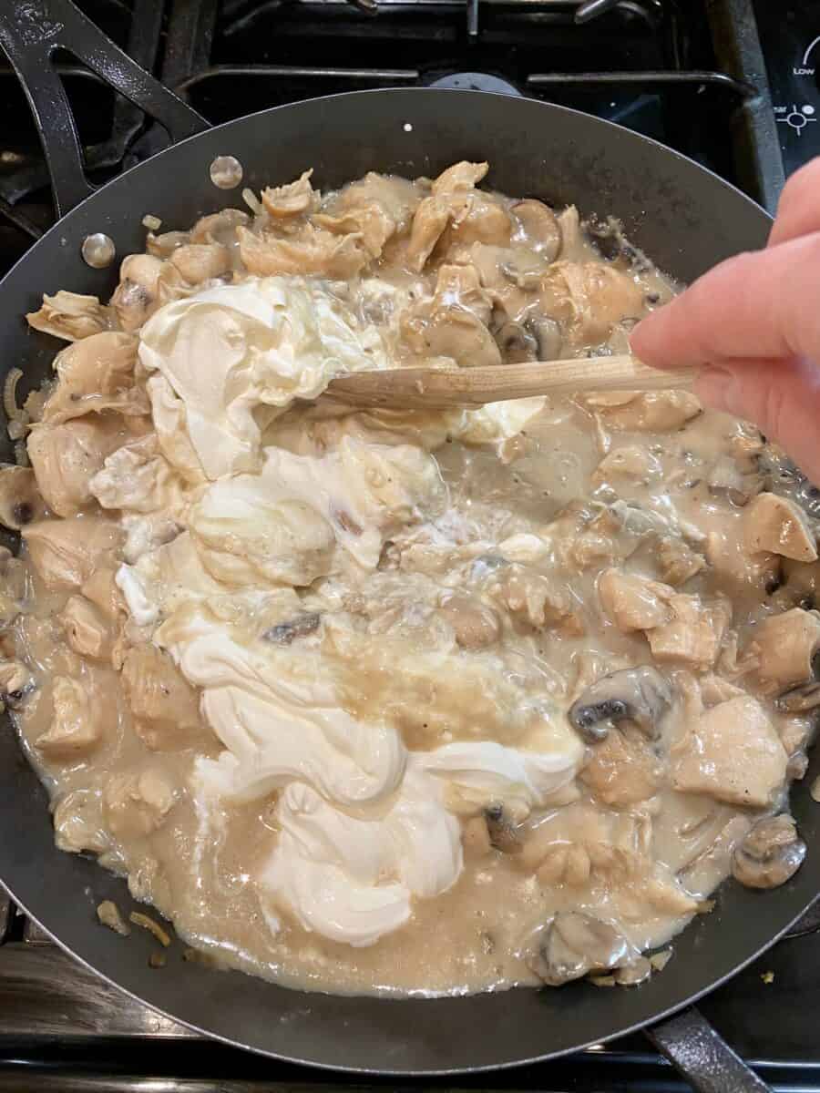 Stirring sour cream into the chicken stroganoff mixture in a skillet on the stove