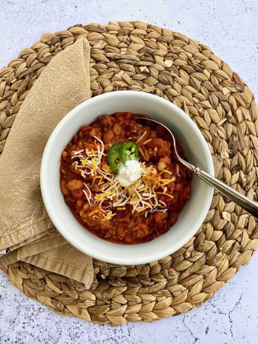 A bowl of chili on a placemat with a napkin on the side of the bowl and a spoon in the chili