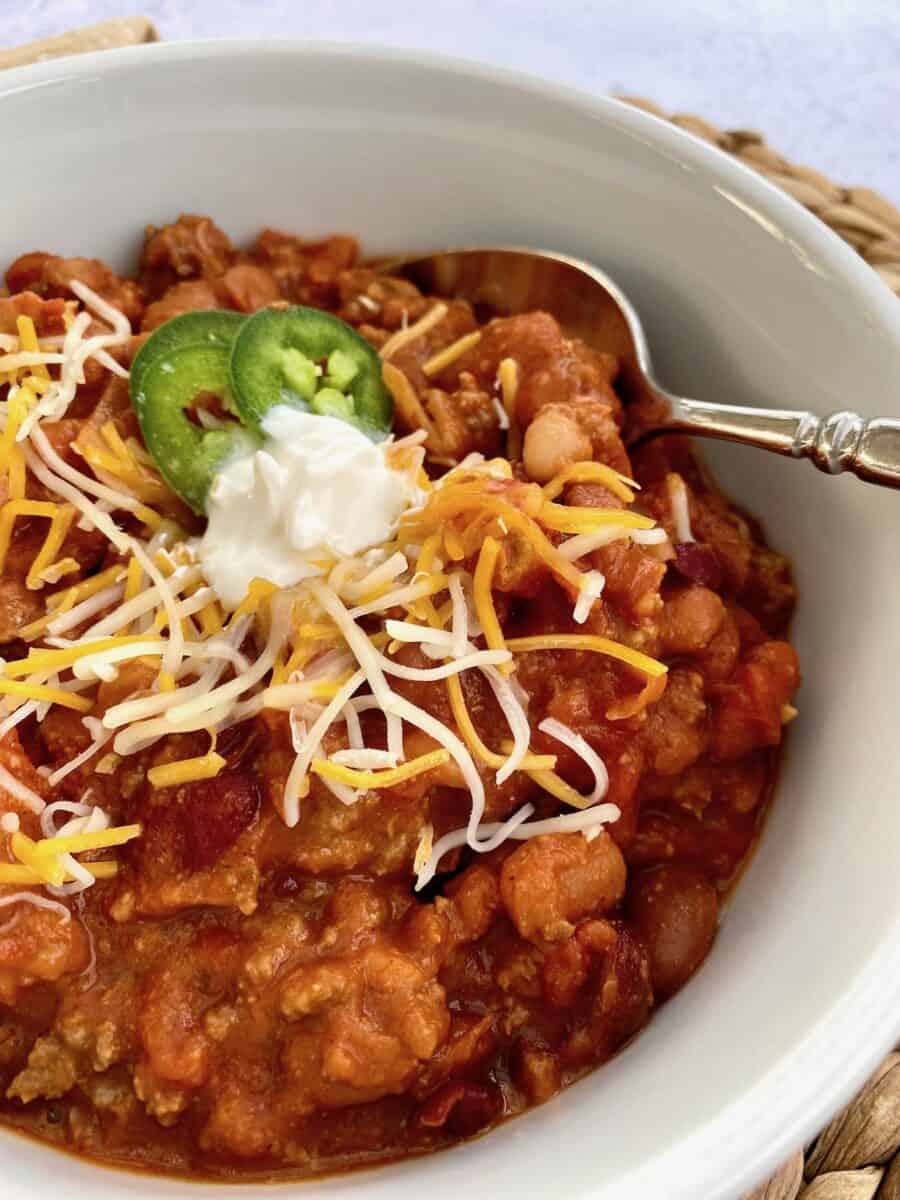 A bowl of chili with shredded cheese, sour cream and jalapenos on top with a spoon on the side