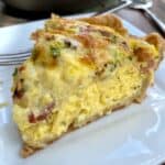 A wedge of quiche on a plate with the quiche dish in the background.