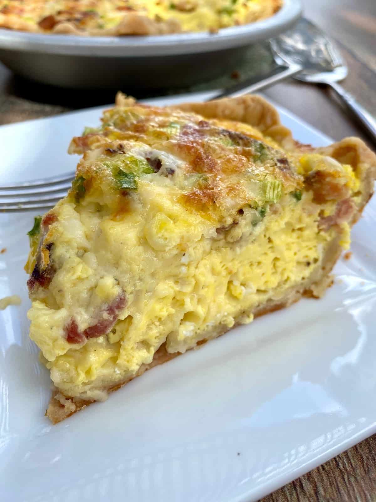 A wedge of quiche on a plate with the quiche dish in the background. 