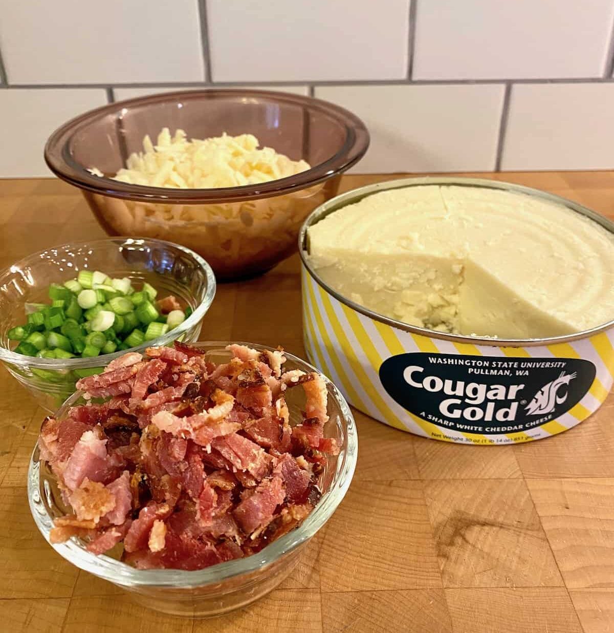 Small bowls of cooked, diced bacon, green onions and shredded cheese with an opened can of Cougar Gold on the side.