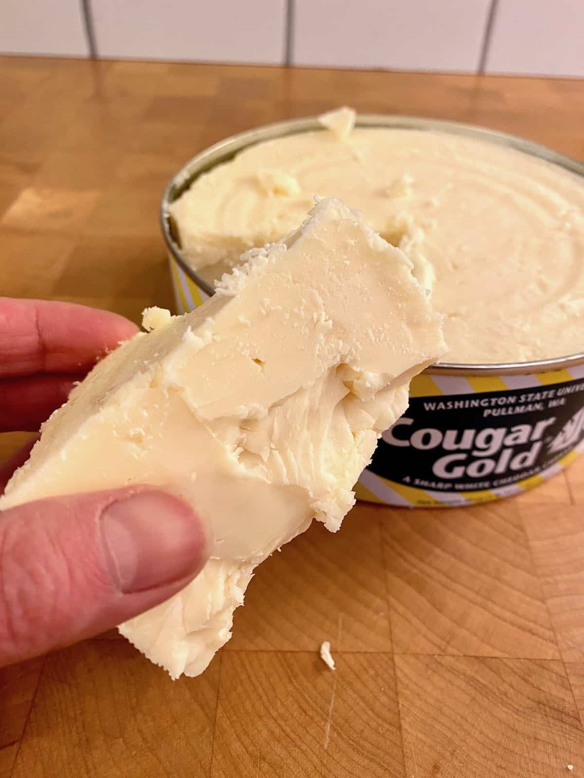 A hand holding a wedge of sharp white cheddar cheese, with the open container of cheese on the side.