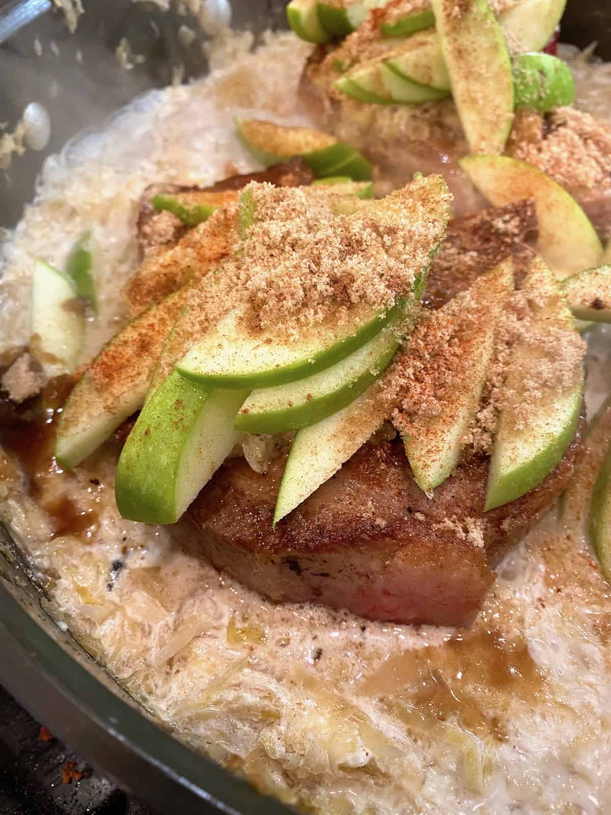 Cooked pork chops topped with sliced apples and spices in a skillet.