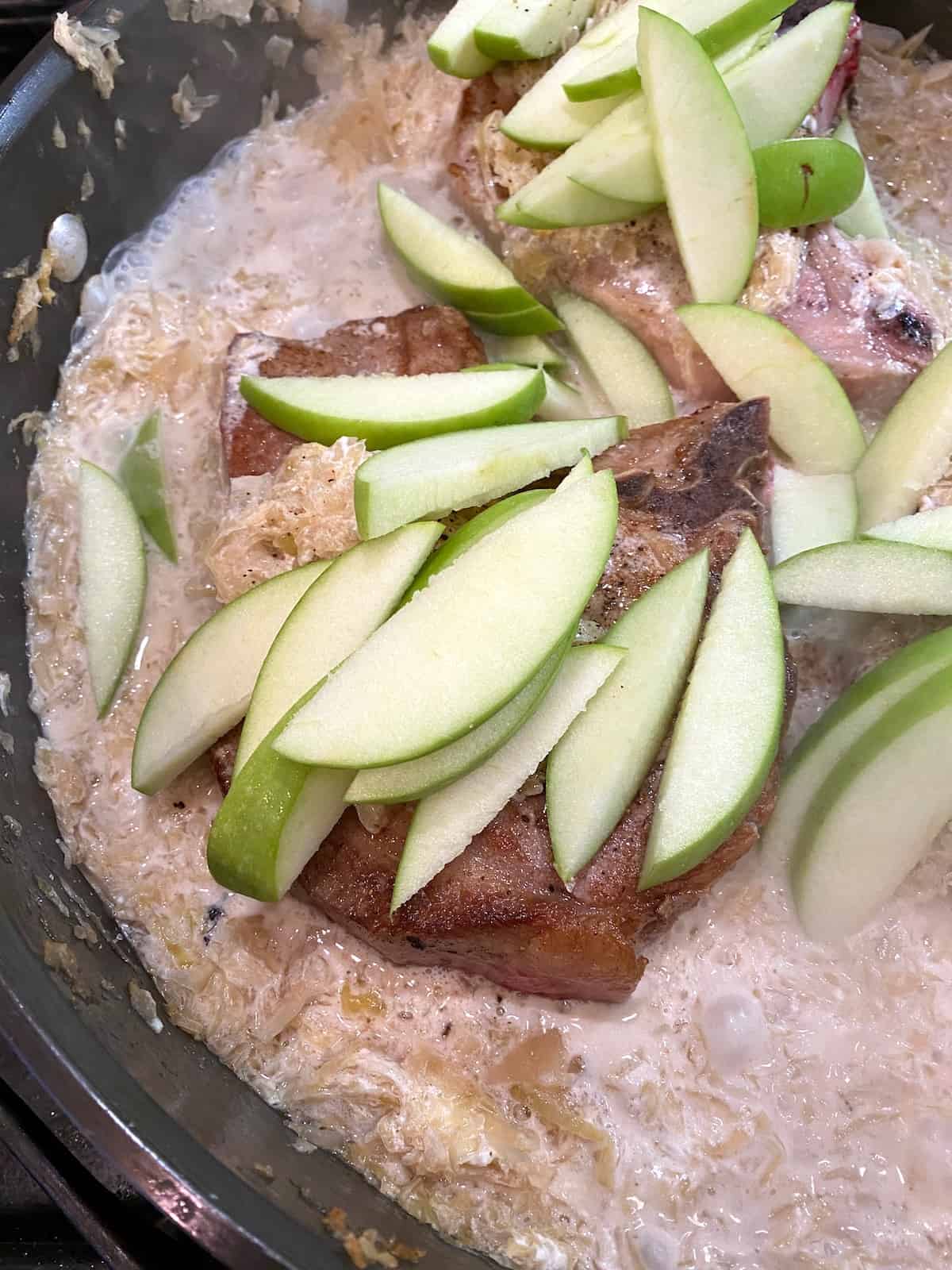 Sliced apples on top of pork chops in a skillet on the stove.