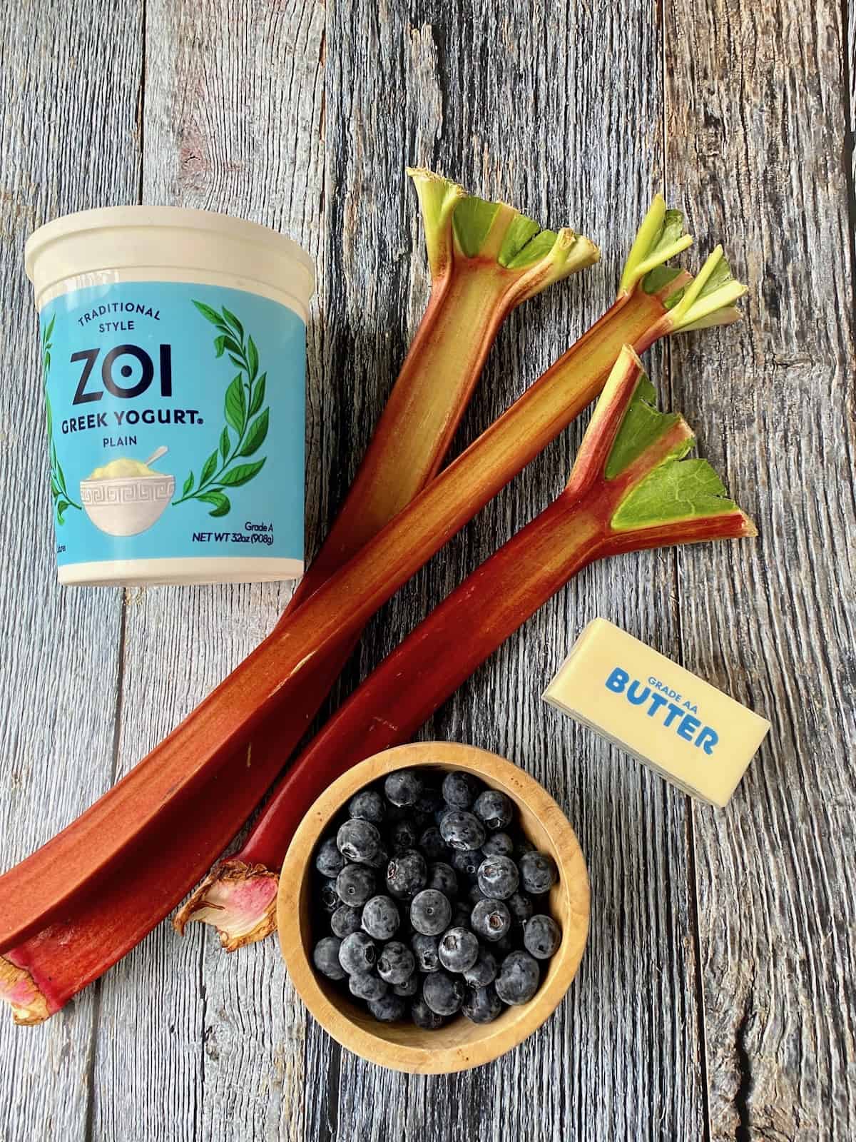 A container of yogurt next to several stalks of rhubarb, a stick of butter and a bowl of blueberries. 