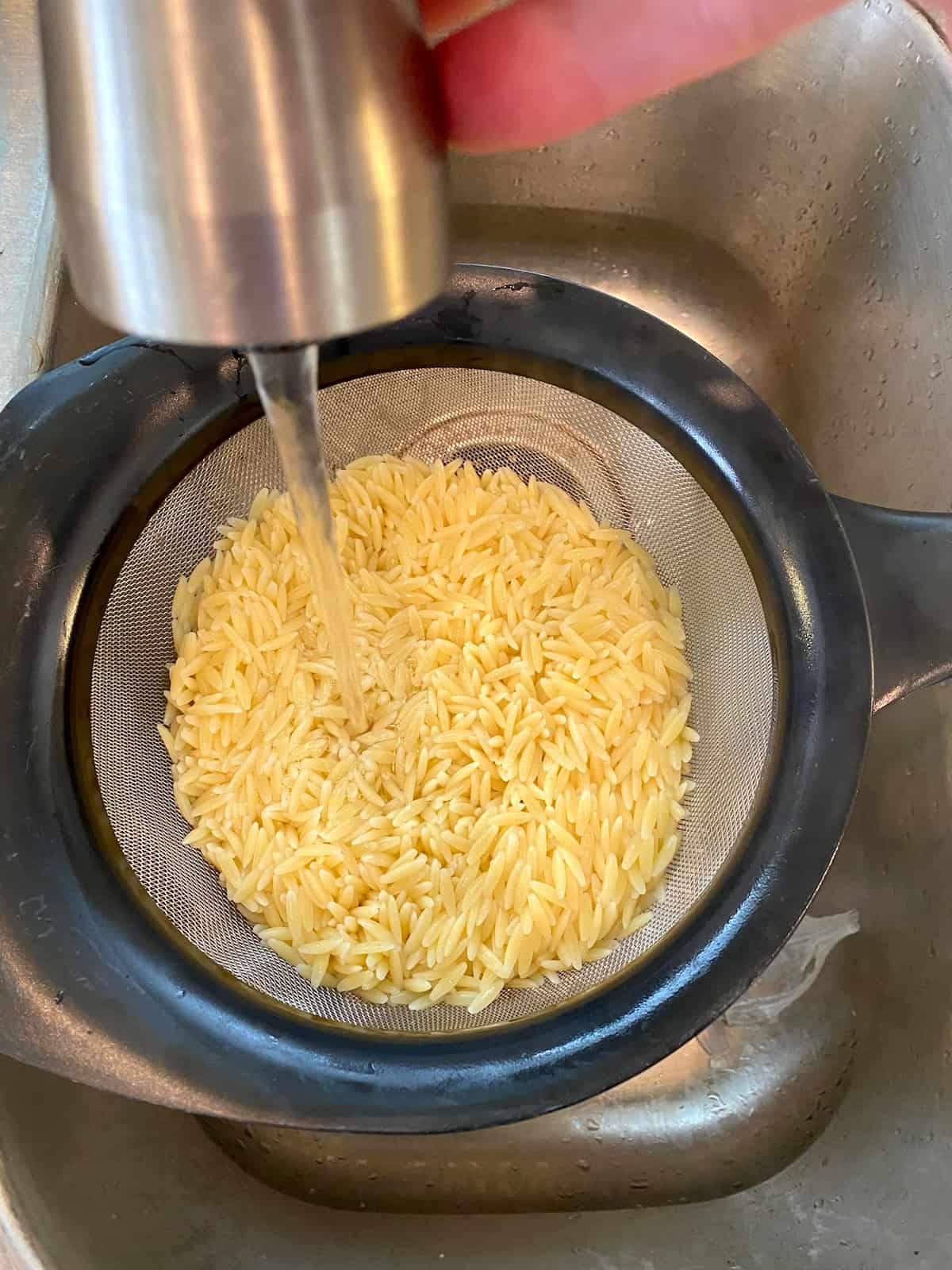 Orzo pasta in a strainer in a sink being rinsed with cool water from a faucet.