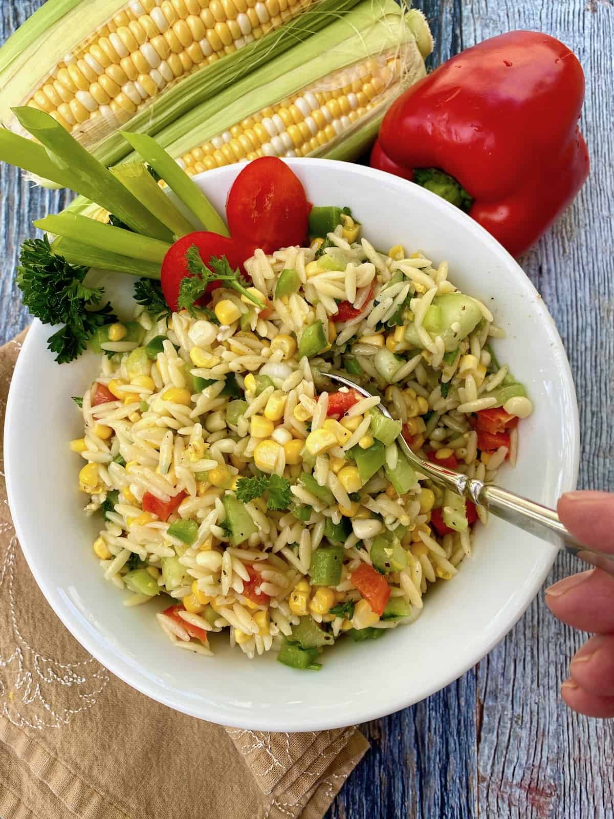 A spoon serving a portion of sweet corn pasta salad from a bowl with several ears of corn and a red pepper in the background.