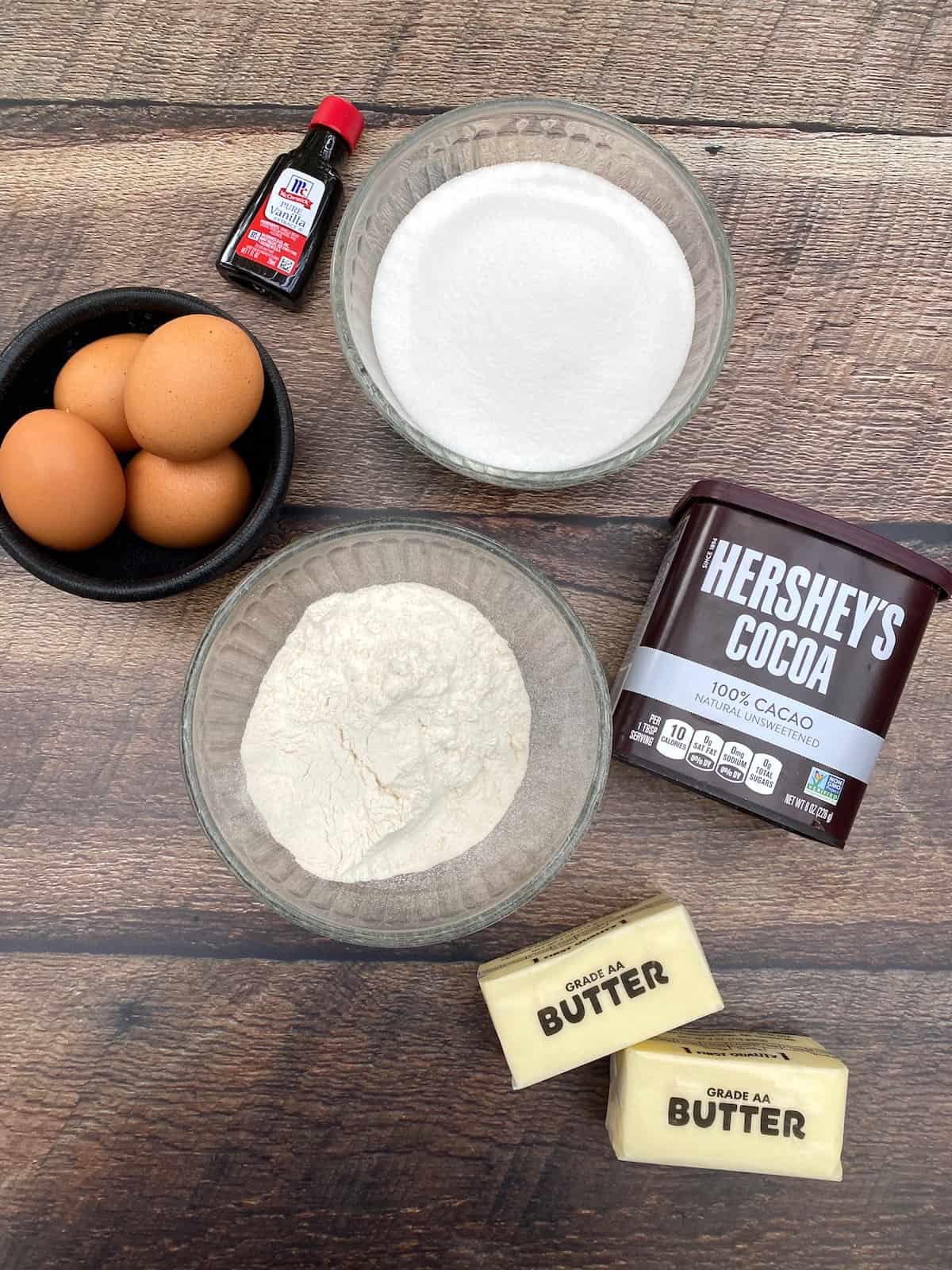 Eggs, vanilla extract, flour, sugar, cocoa powder and two sticks of butter displayed on a plank.