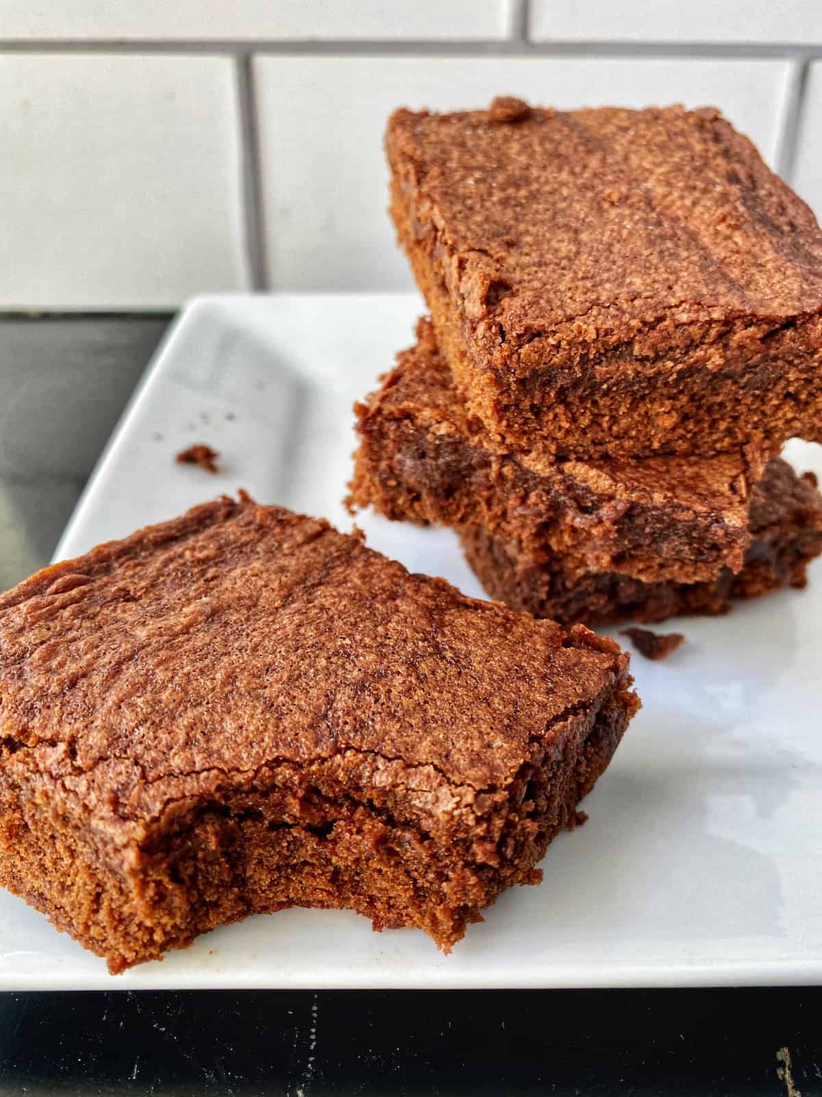 Three brownies stacked on a plate, one in front with a bite taken out of it.