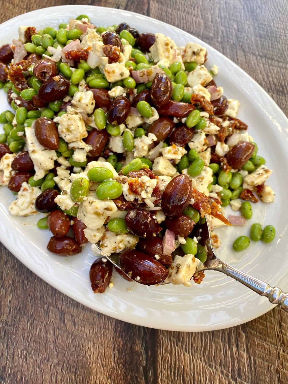 A large plate with mediterranean edamame salad spread out on it, with a spoon scooping up one serving.