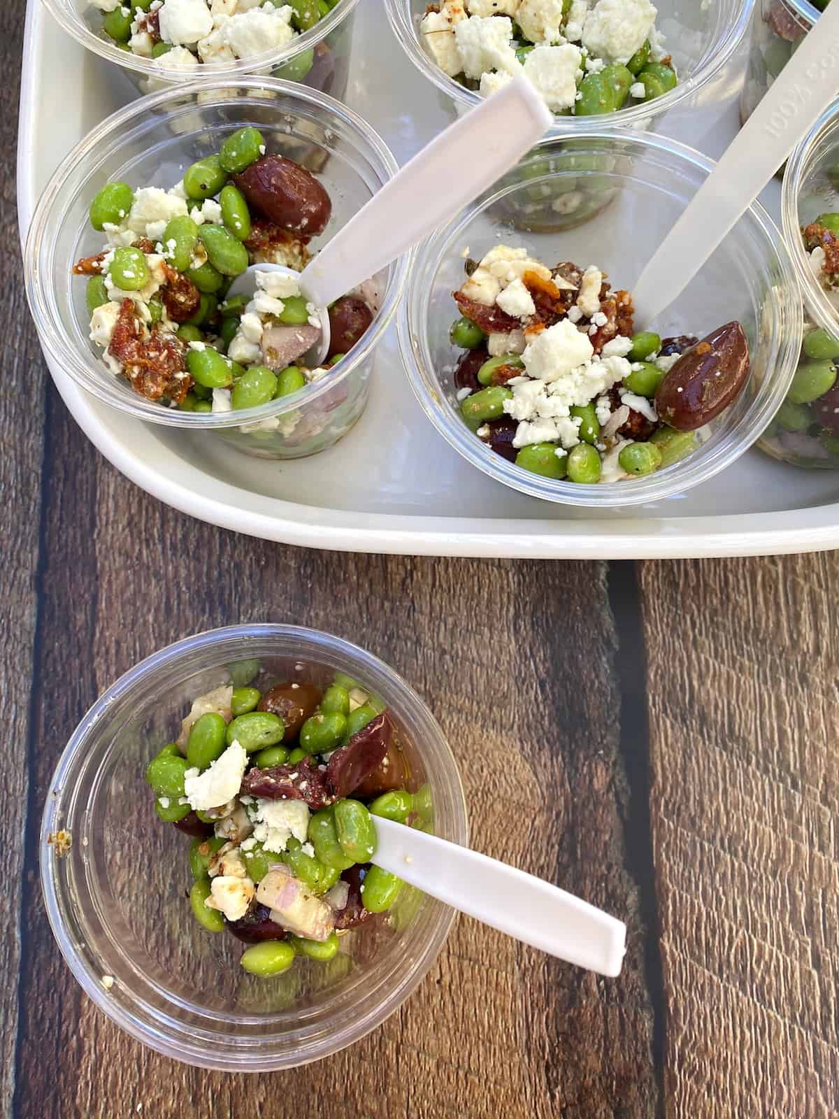 Small cups of salad with a spoon in each one, displayed in a serving dish with one small cup off to the side.