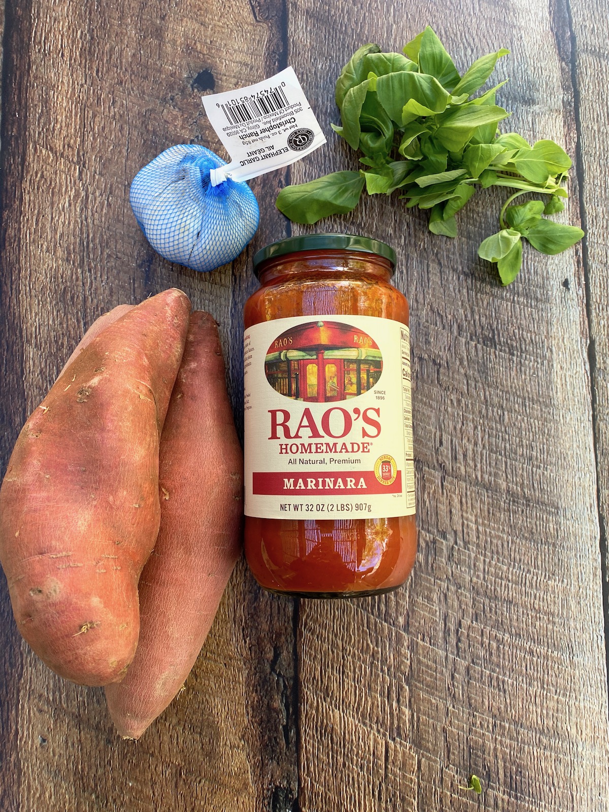 Ingredients arranged on a wooden plank, sweet potatoes, marinara sauce in a jar, fresh basil and a bulb of elephant garlic. 