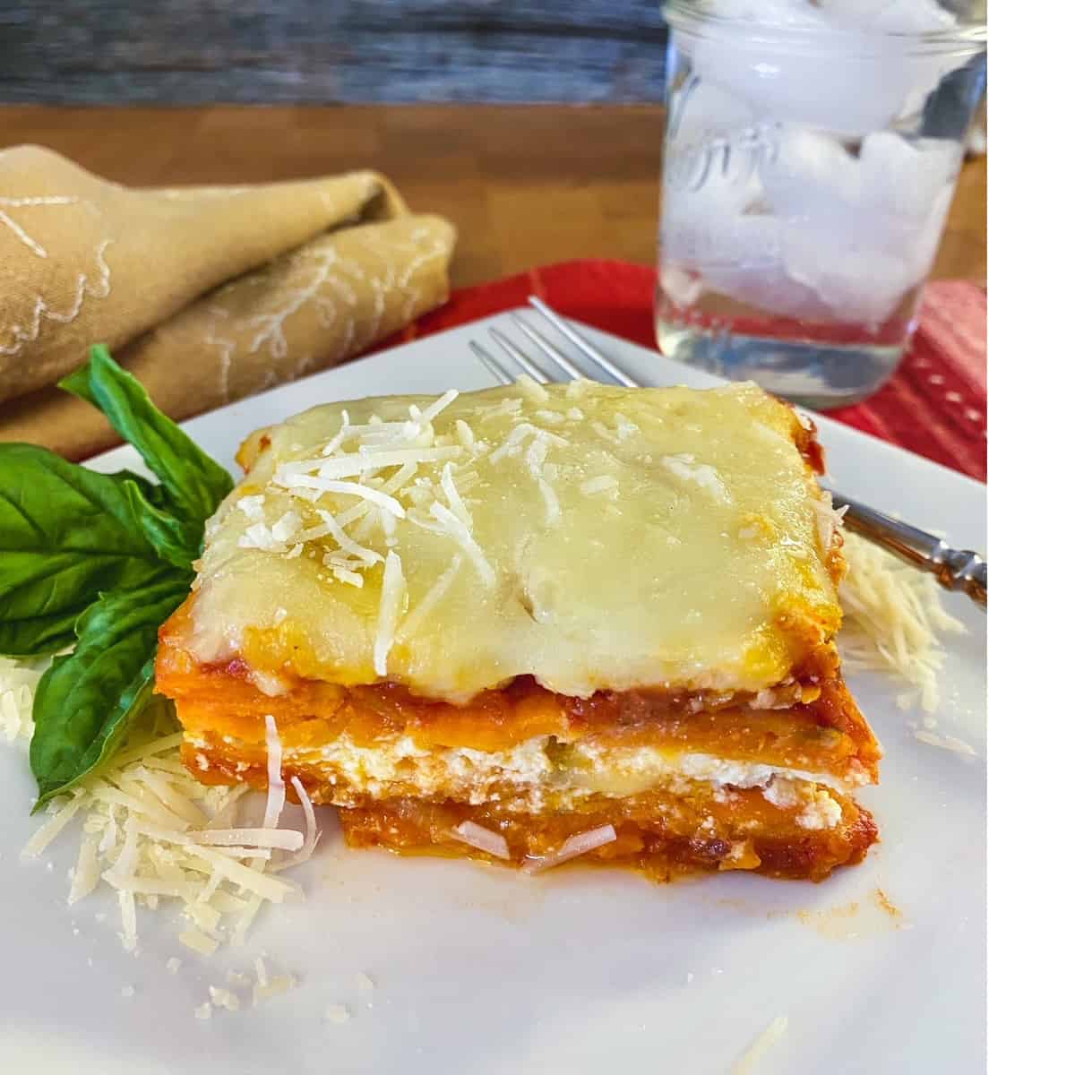 A slice of sweet potato lasagna on a plate with a glass of ice water on the side.