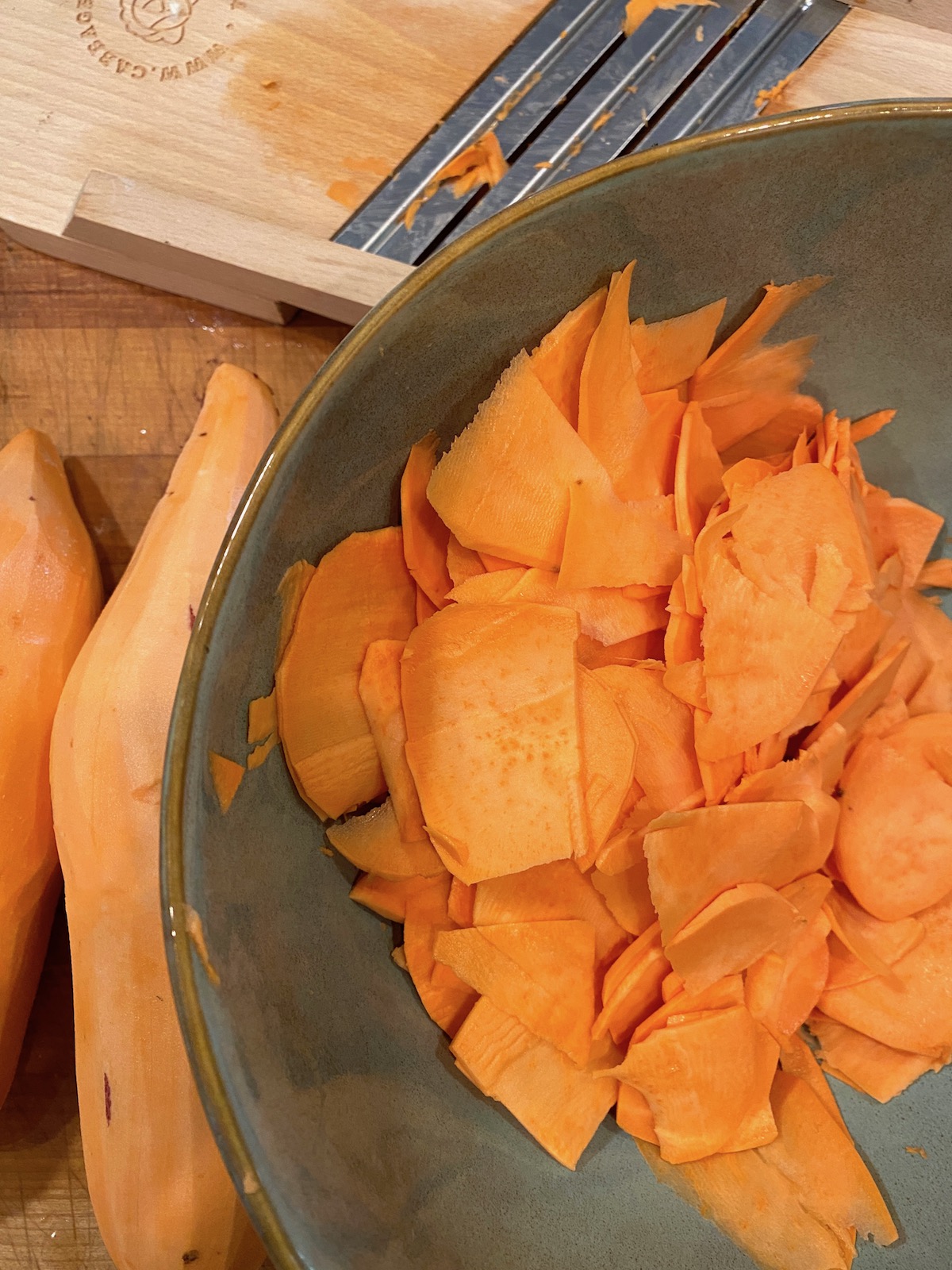 A bowl of thinly sliced sweet potatoes with several peeled sweet potatoes on the side. 