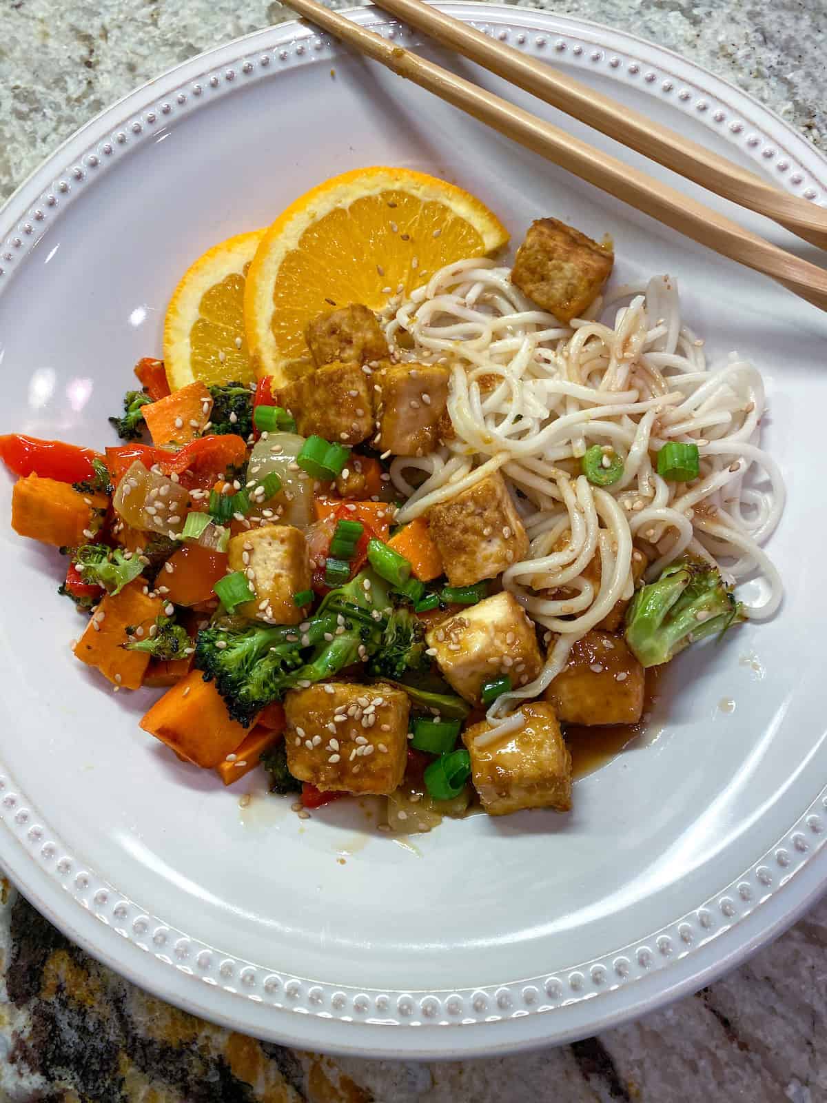 A plate of baked tofu chunks with vegetables and noodles with orange glaze on top.