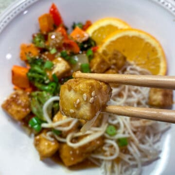 A plate of orange tofu with veggies and noodles with orange slices on the side.