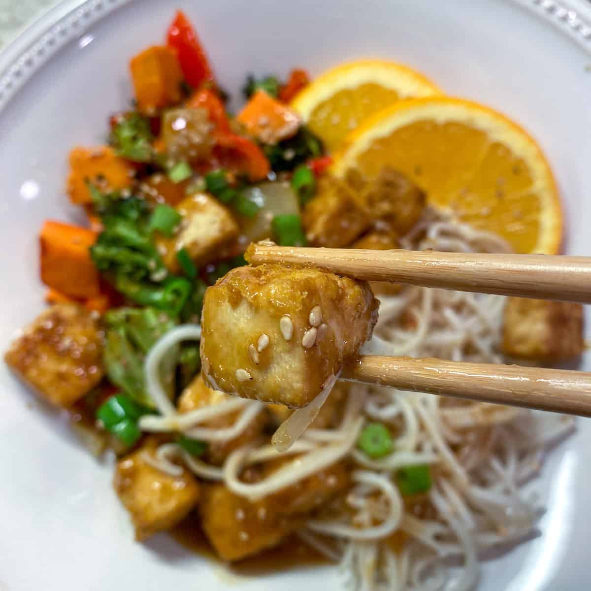 A plate of orange tofu with veggies and noodles with orange slices on the side. 