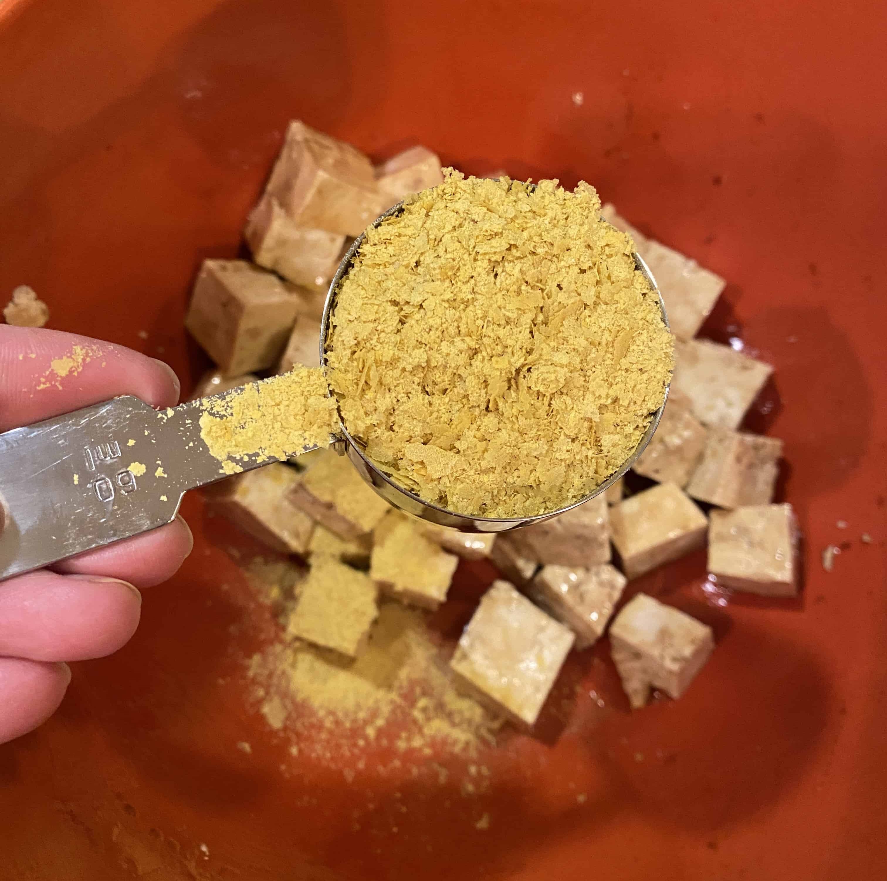 A scoop of nutritional yeast being held over a bowl of diced tofu.